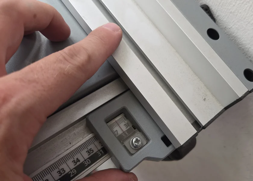 Ein Finger zeigt auf die integrierte Skala mit Ableselupe am silbernen Parallelanschlag der Tischkreissäge.
